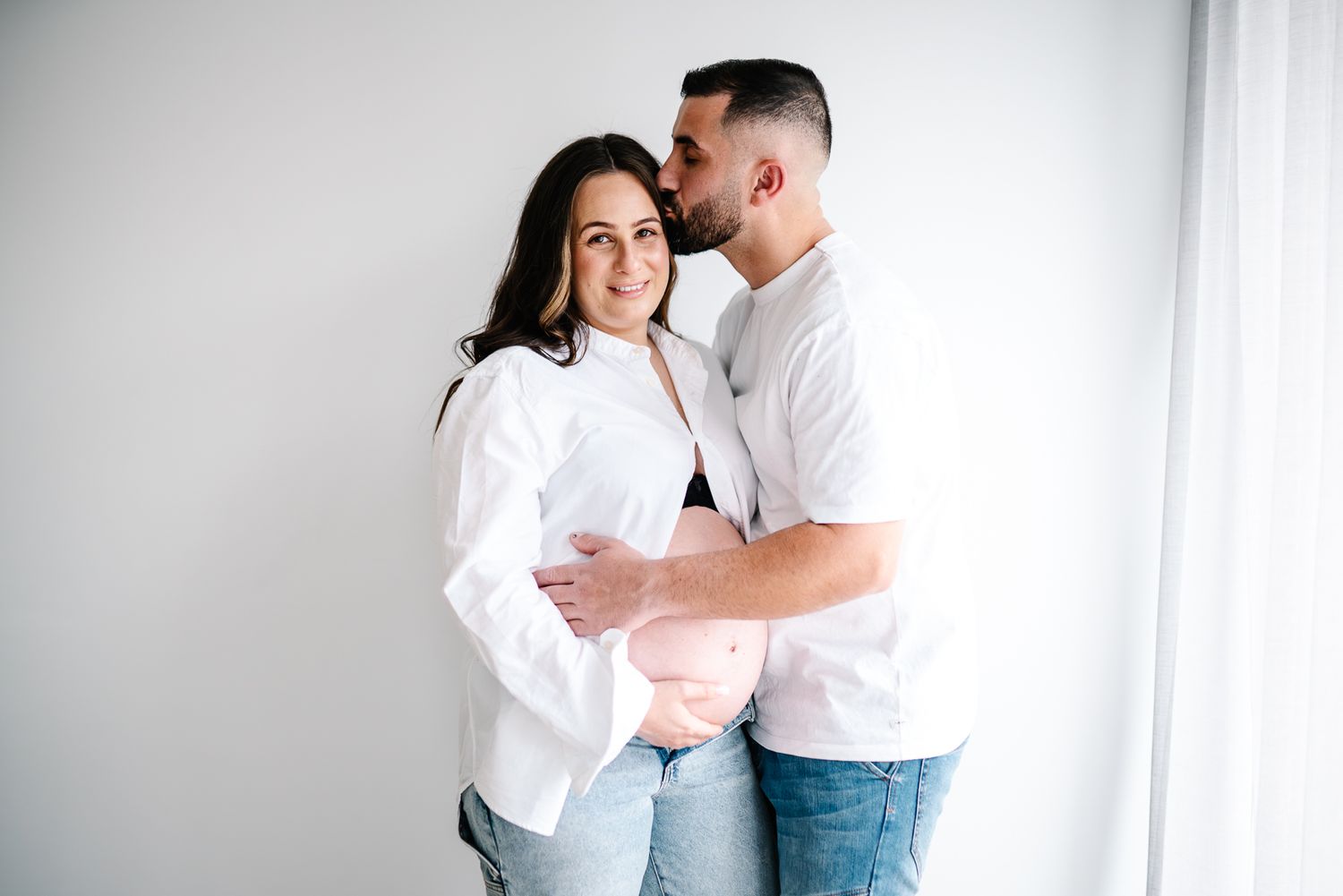 A pregnant couple in white outfits shares an intimate moment against a bright window backdrop during a maternity photo session in Melbourne.