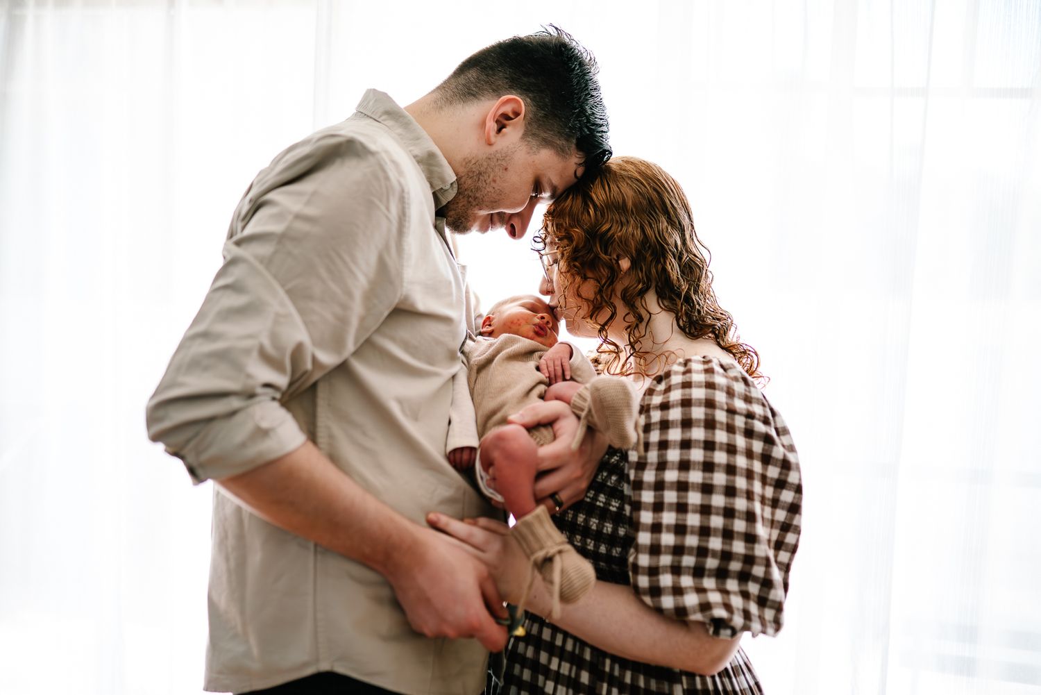 A joyful family moment captured indoors against white curtains as they embrace together during a Melbourne newborn and family photo session.