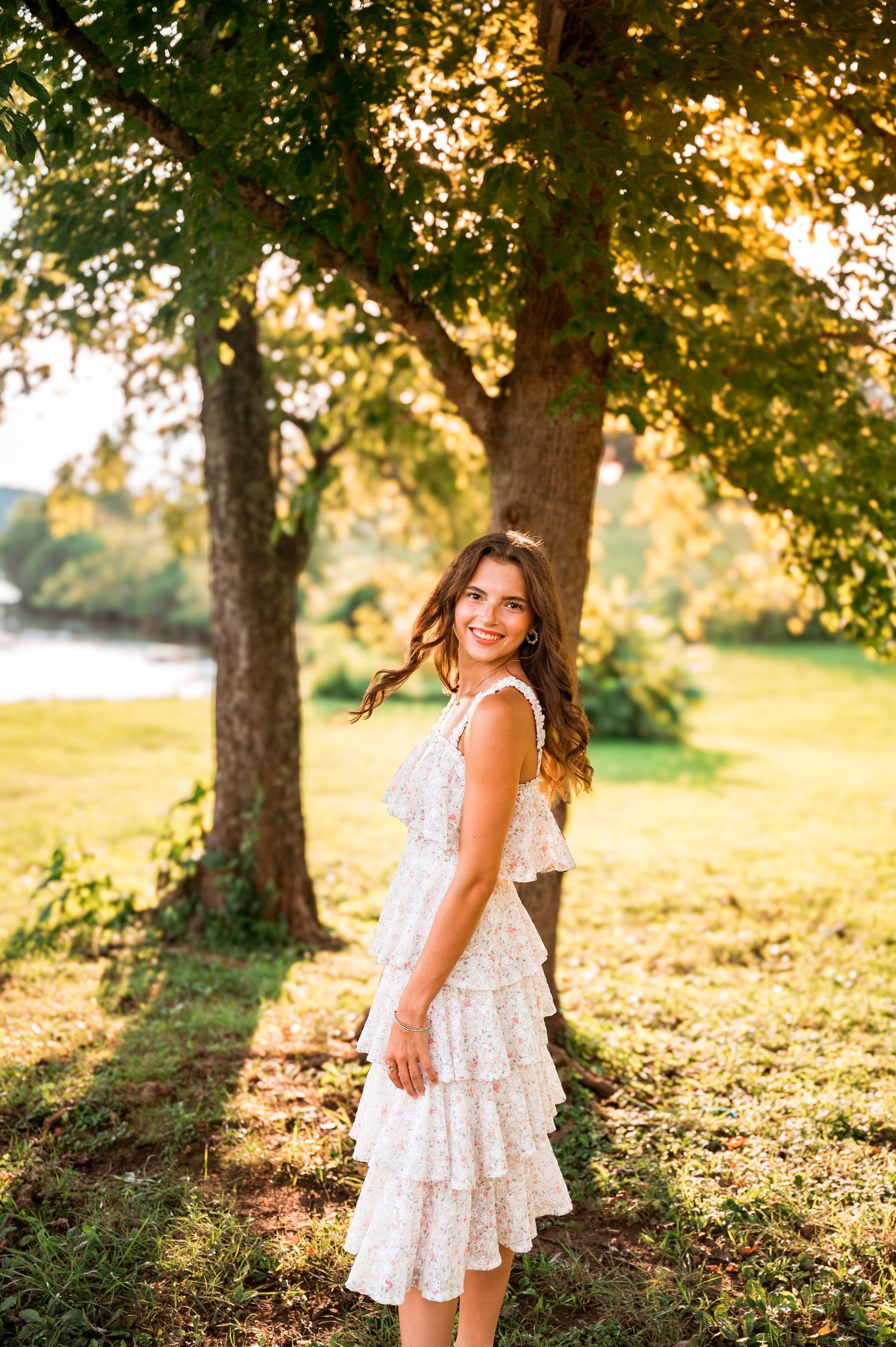 A white lace dress twirls in golden sunlight beneath green trees during a summer photoshoot.