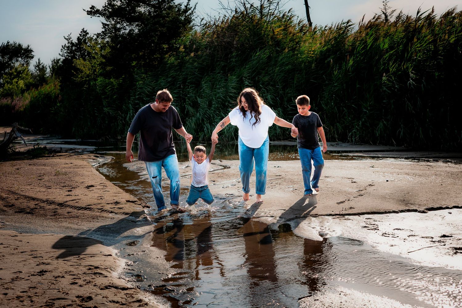 A group walks along a beach at sunset, reflecting in wet sand as they hold hands while walking near tall grass.