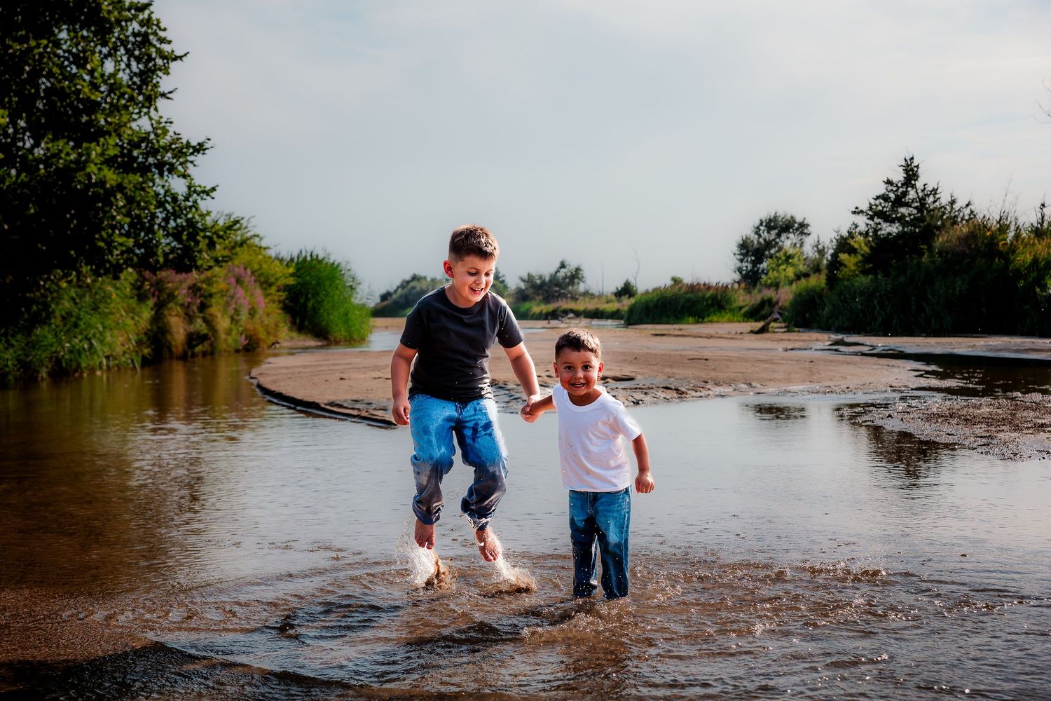 Two boys splashing through a muddy puddle outdoors, laughing and running together on a sunny day.