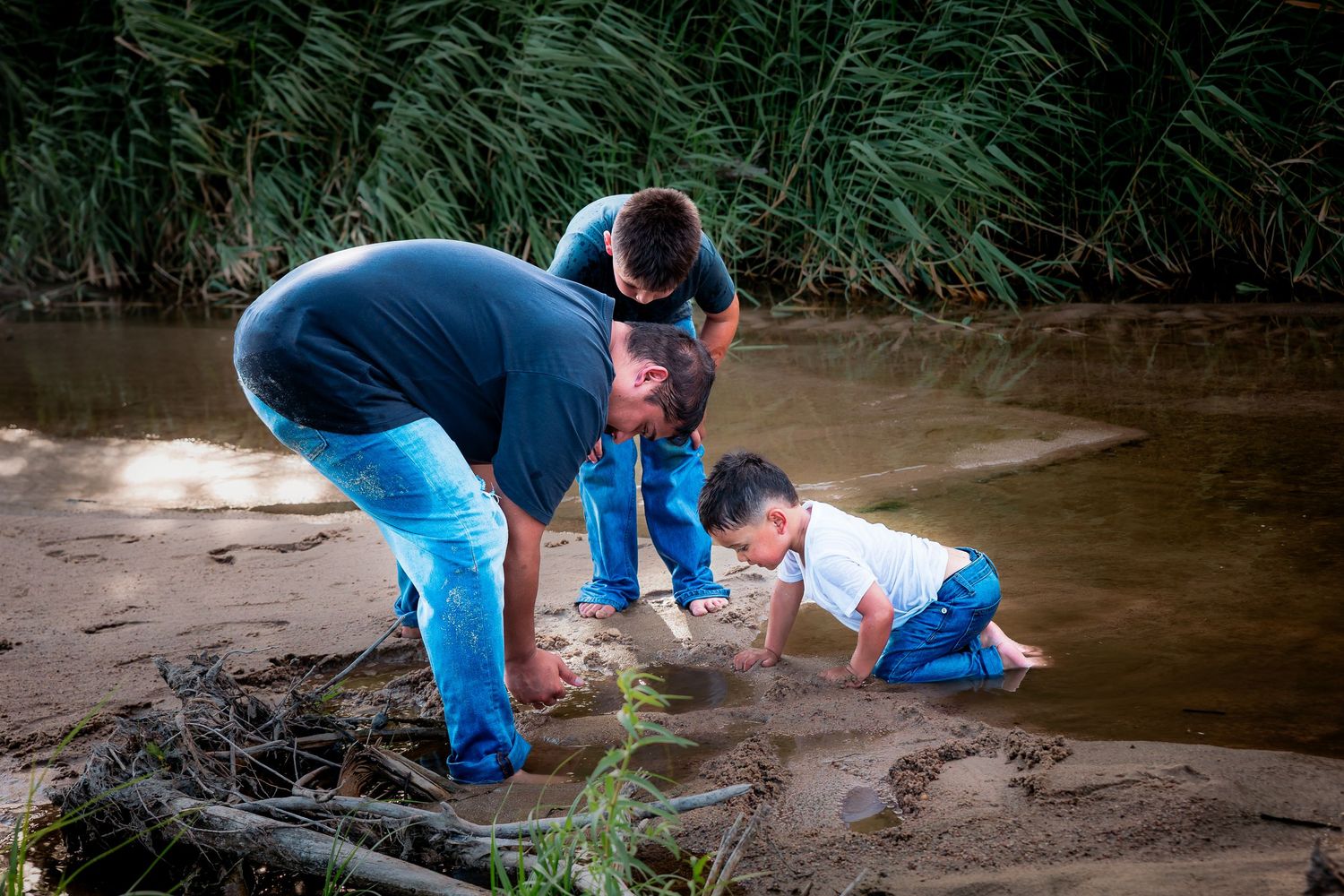 A father and two young children explore a muddy riverbank, examining the water's edge surrounded by tall green reeds.