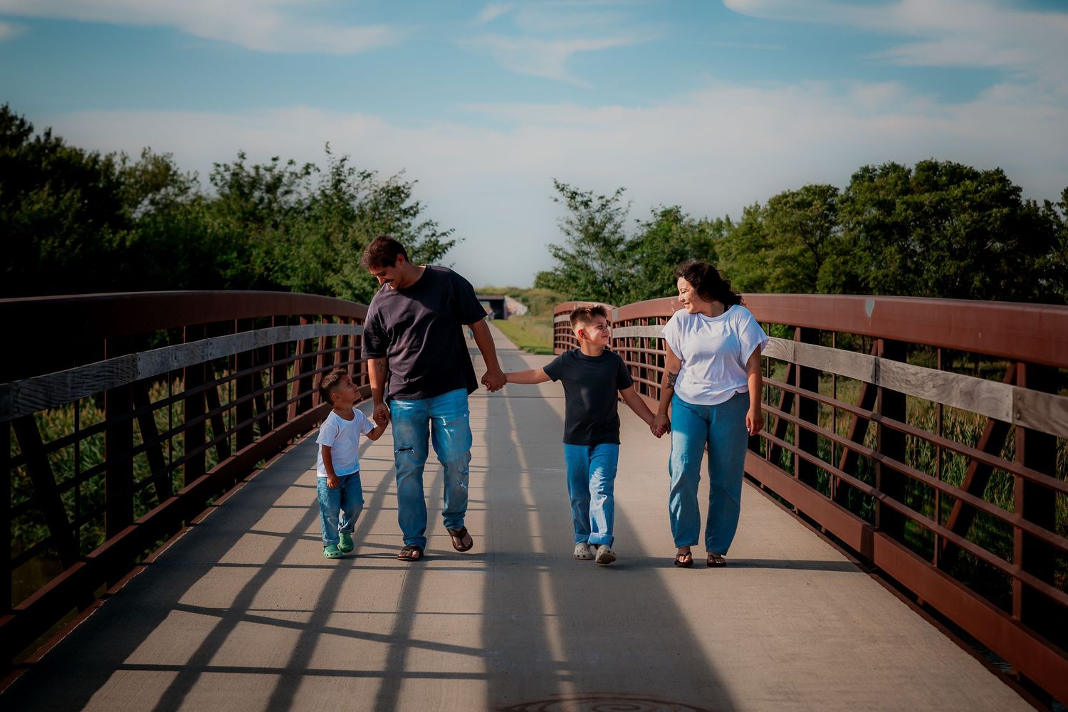 Family of four holding hands while walking together on a wooden bridge surrounded by green trees.