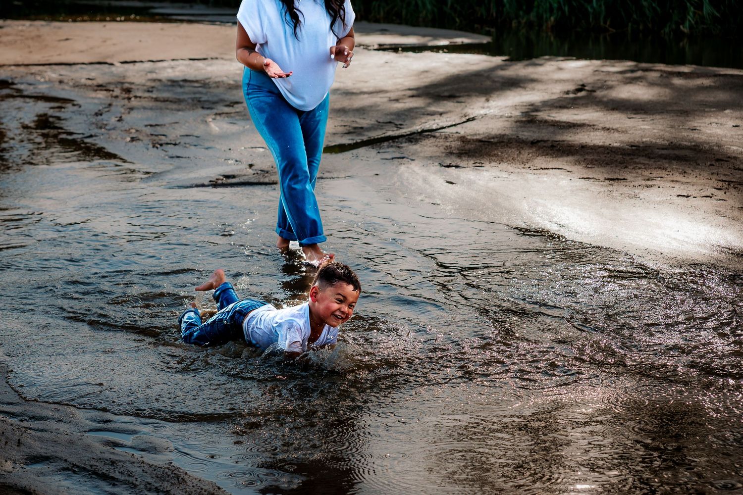 Child playing in shallow water while adult stands nearby on wet ground outdoors.