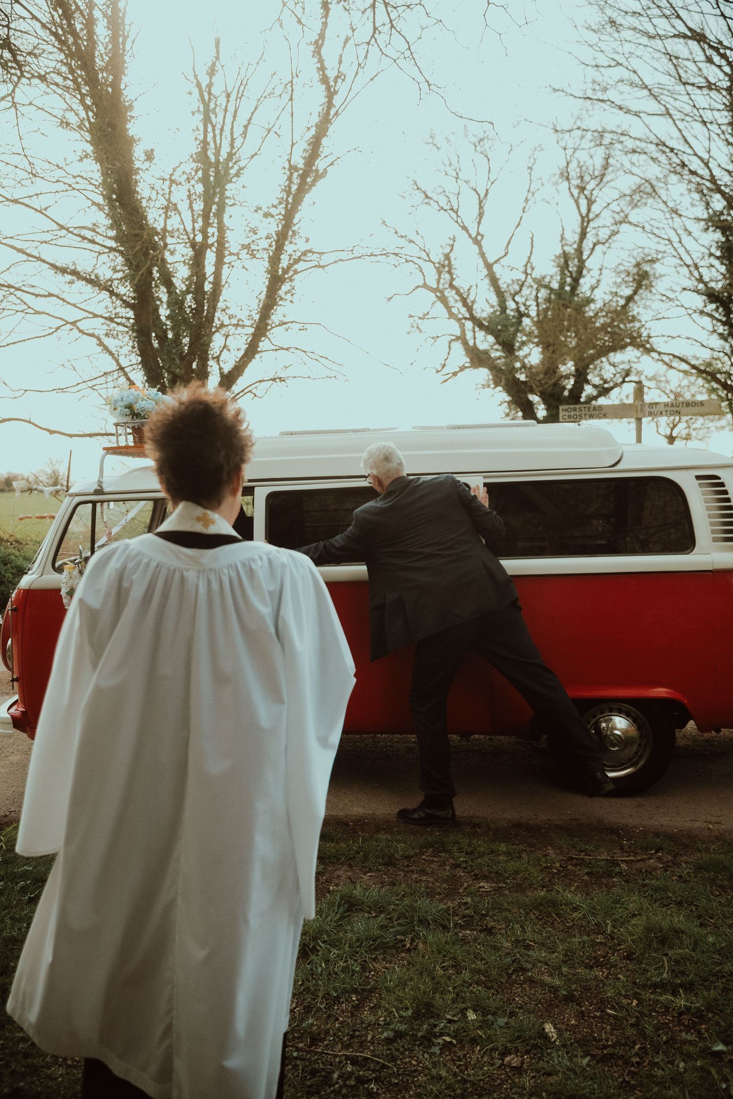 Vicor in white robes watches a bride's father opening the sliding door to a vintage red and white VW camper van to help out a bride.