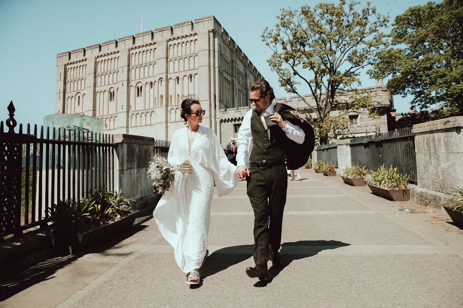 A couple in wedding attire walks along a stone wall pathway on a sunny day.