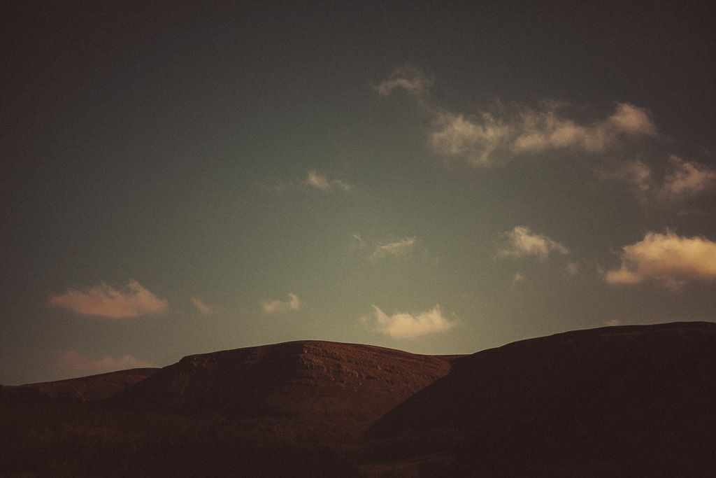 Dark moody landscape with rolling hills silhouetted against a teal sky with scattered clouds at dusk in Wales.