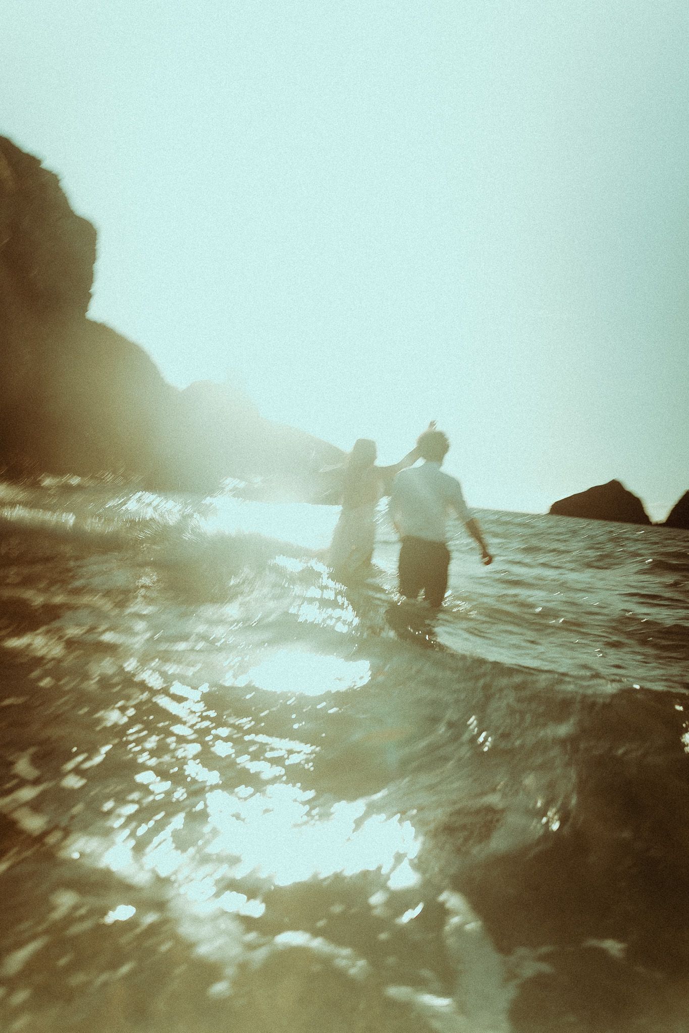 Couple wading through ocean waves near rocky shore in dreamy, blurred vintage-style beach photography.