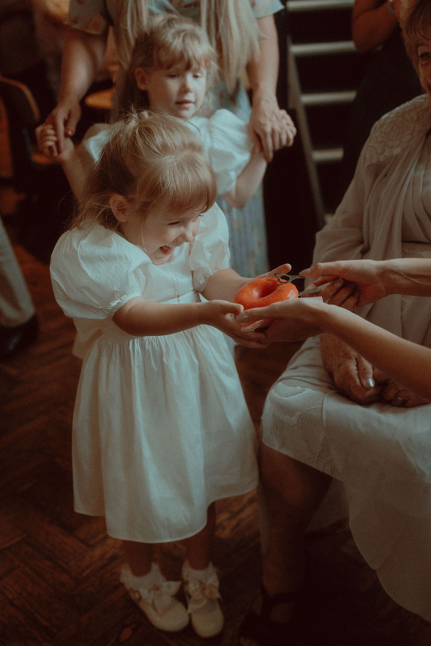 Young flower girl in white dress handing a small orange gift to a seated woman at a wedding ceremony.