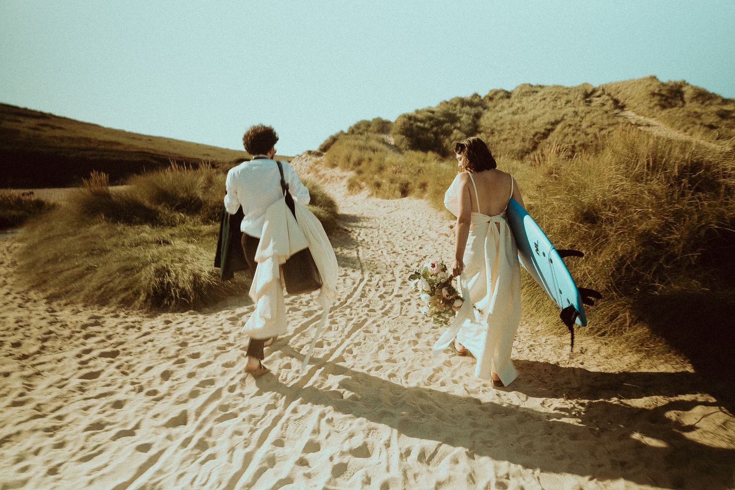 Two people in white outfits walk barefoot along a sandy dune path, one carrying a bouquet and the other a blue surfboard, in golden light.