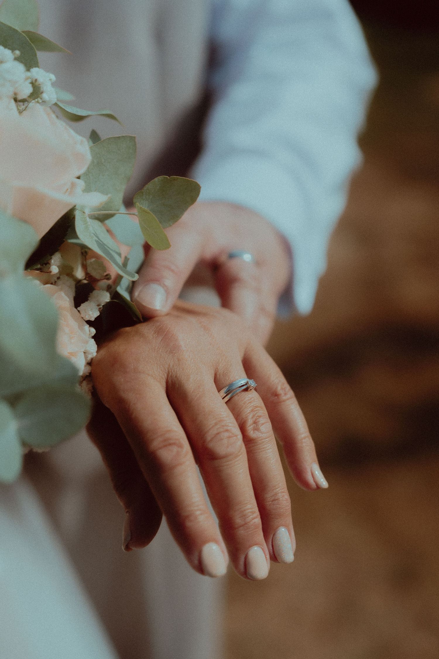 Close-up of two hands displaying wedding rings, holding a bouquet of white flowers and eucalyptus leaves.