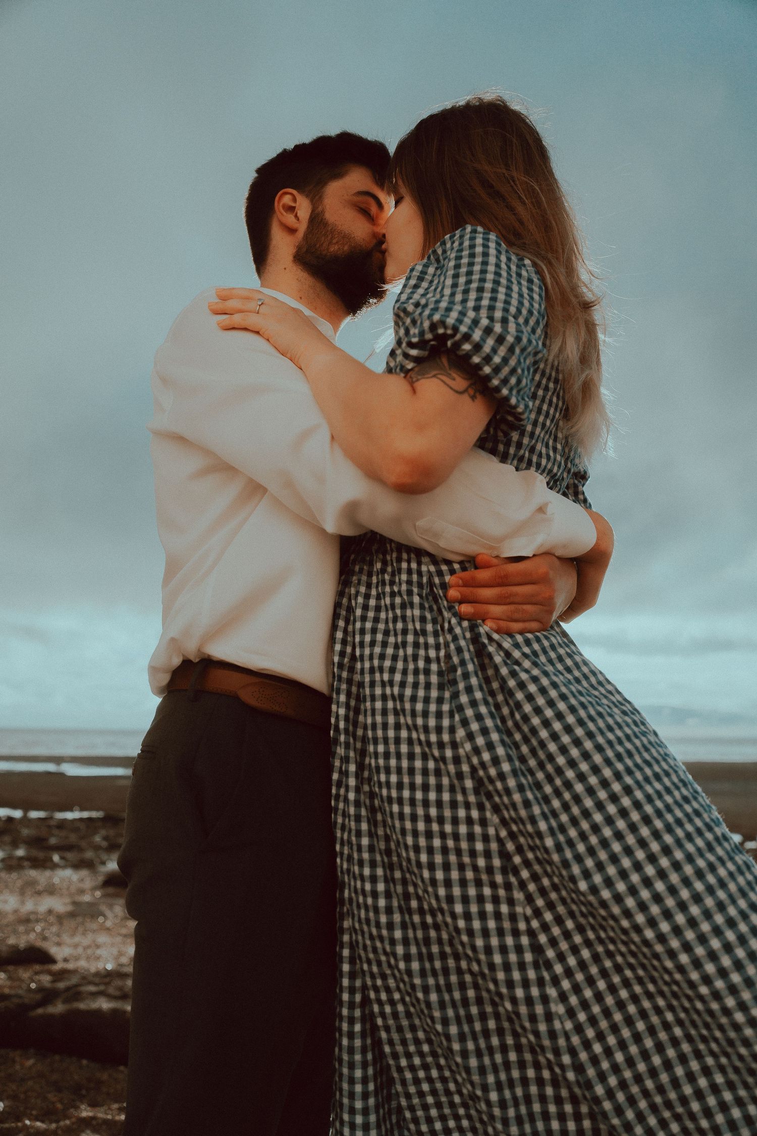 A couple sharing a romantic kiss while embracing outdoors near a beach, woman in plaid dress, man in white shirt.