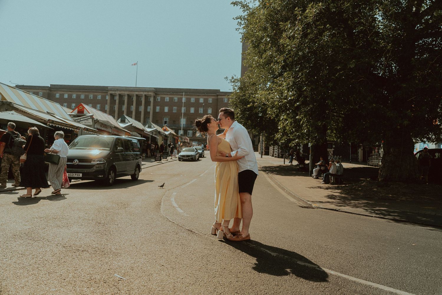 Couple embracing in the middle of a sunlit city street near a bustling outdoor market with tents, parked cars, and tall trees.