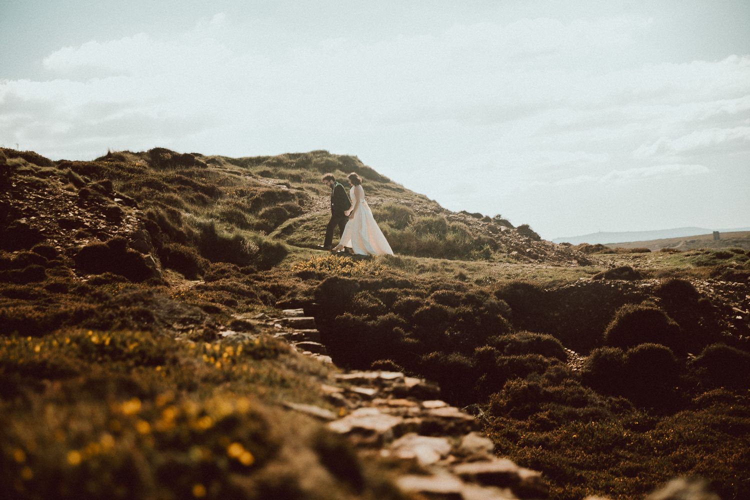 A bride and groom walk through a rugged rocky landscape under a moody overcast sky at sunset.