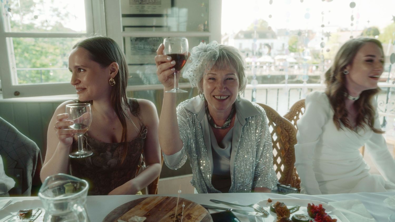 Three women smiling and raising wine glasses in a toast at an elegant dinner table by a bright window.