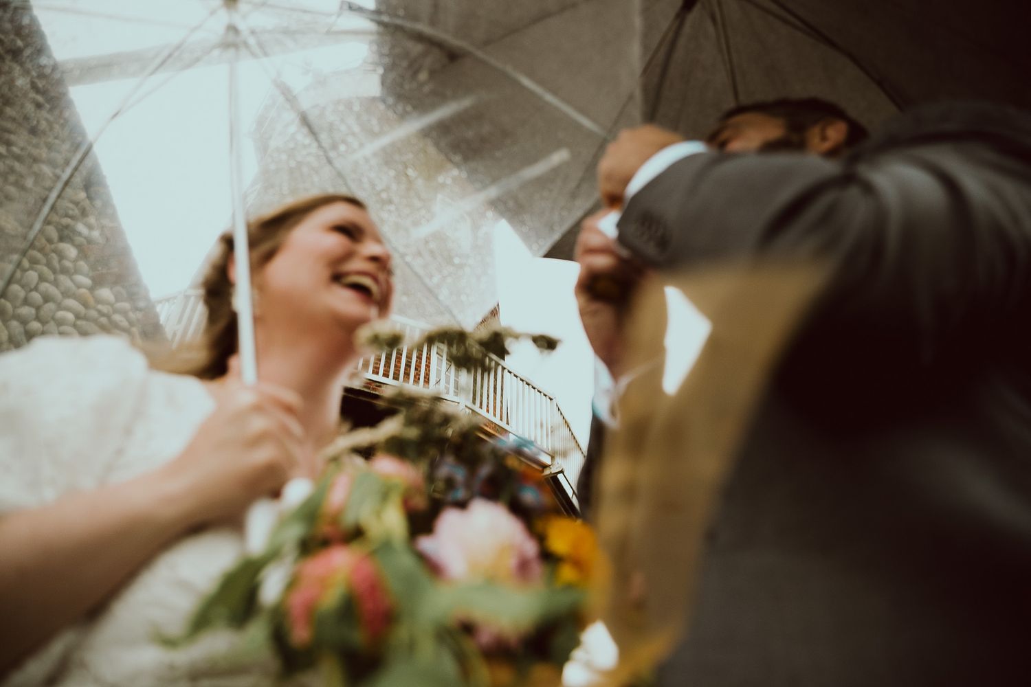 Bride laughing joyfully under a clear umbrella in the rain, holding a colorful bouquet at an outdoor wedding ceremony.