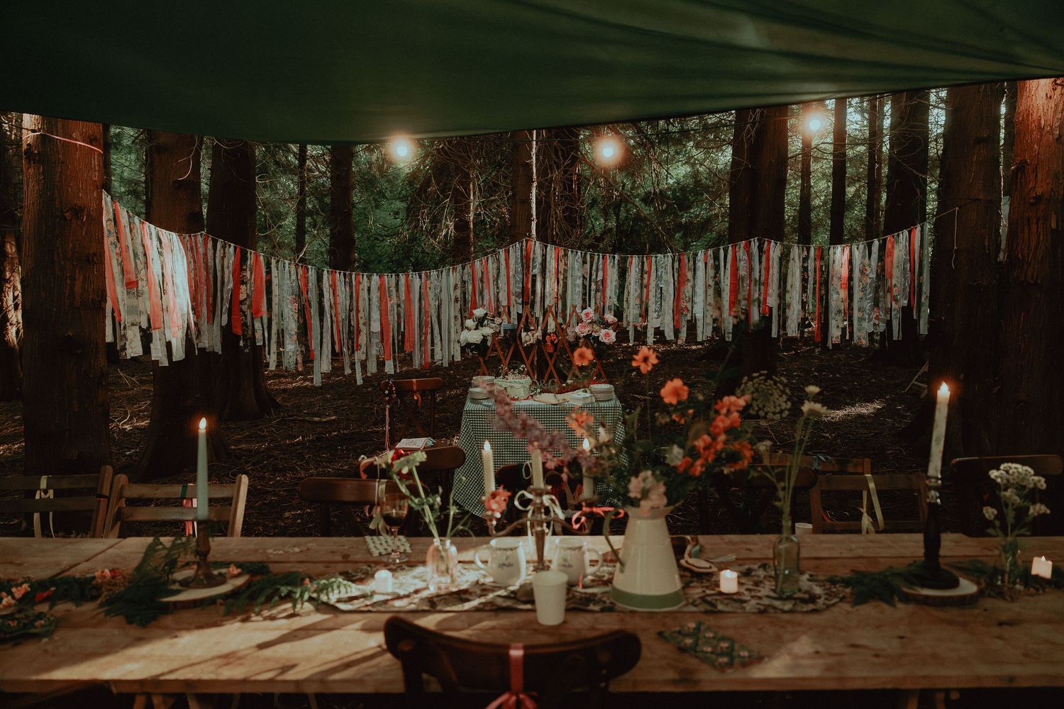 Rustic bohemian wedding reception setup with fabric garland, floral centerpieces, candles, and wooden tables in a forest setting.