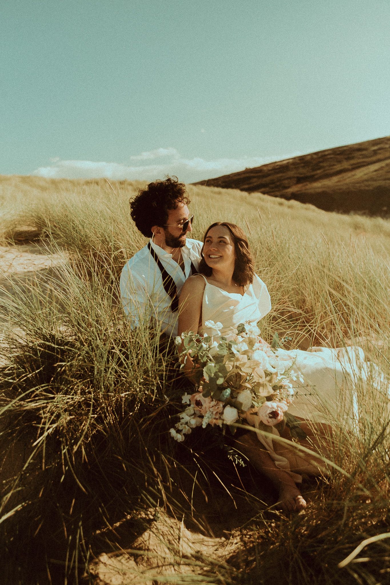 A couple sits together in tall golden grass on a hillside during sunset while holding a white floral bouquet.