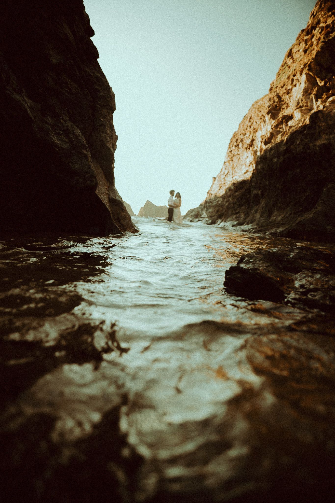 Couple standing between rocky cliffs at the ocean's edge, water flowing in foreground, romantic coastal elopement photo.