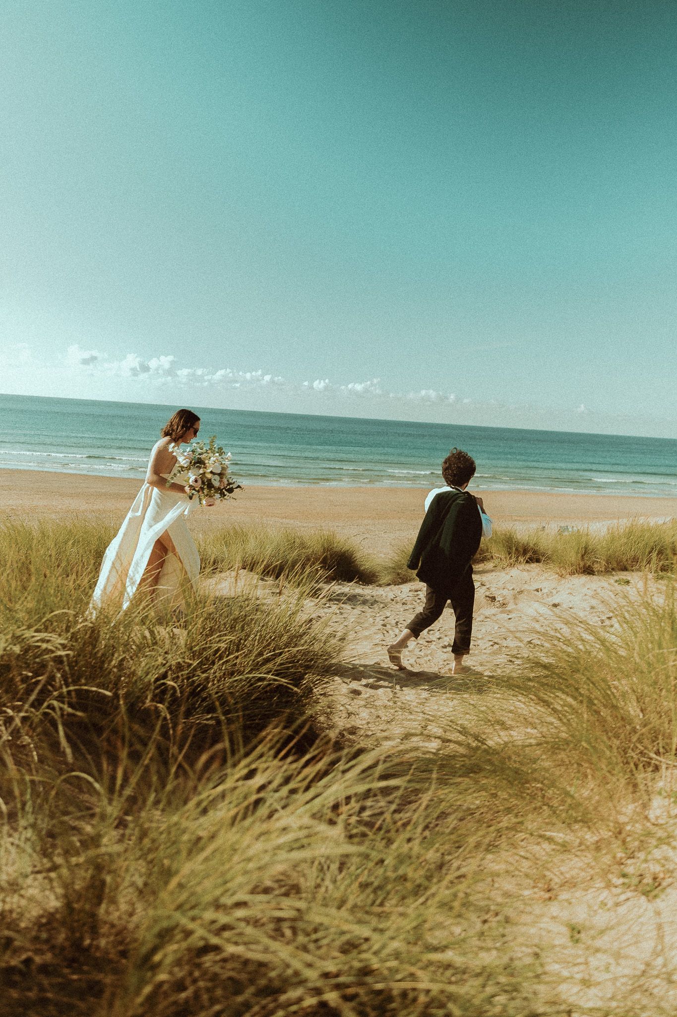 Bride in a flowing white gown holding a bouquet walks toward a groom in a dark suit through beach dunes with the ocean behind.