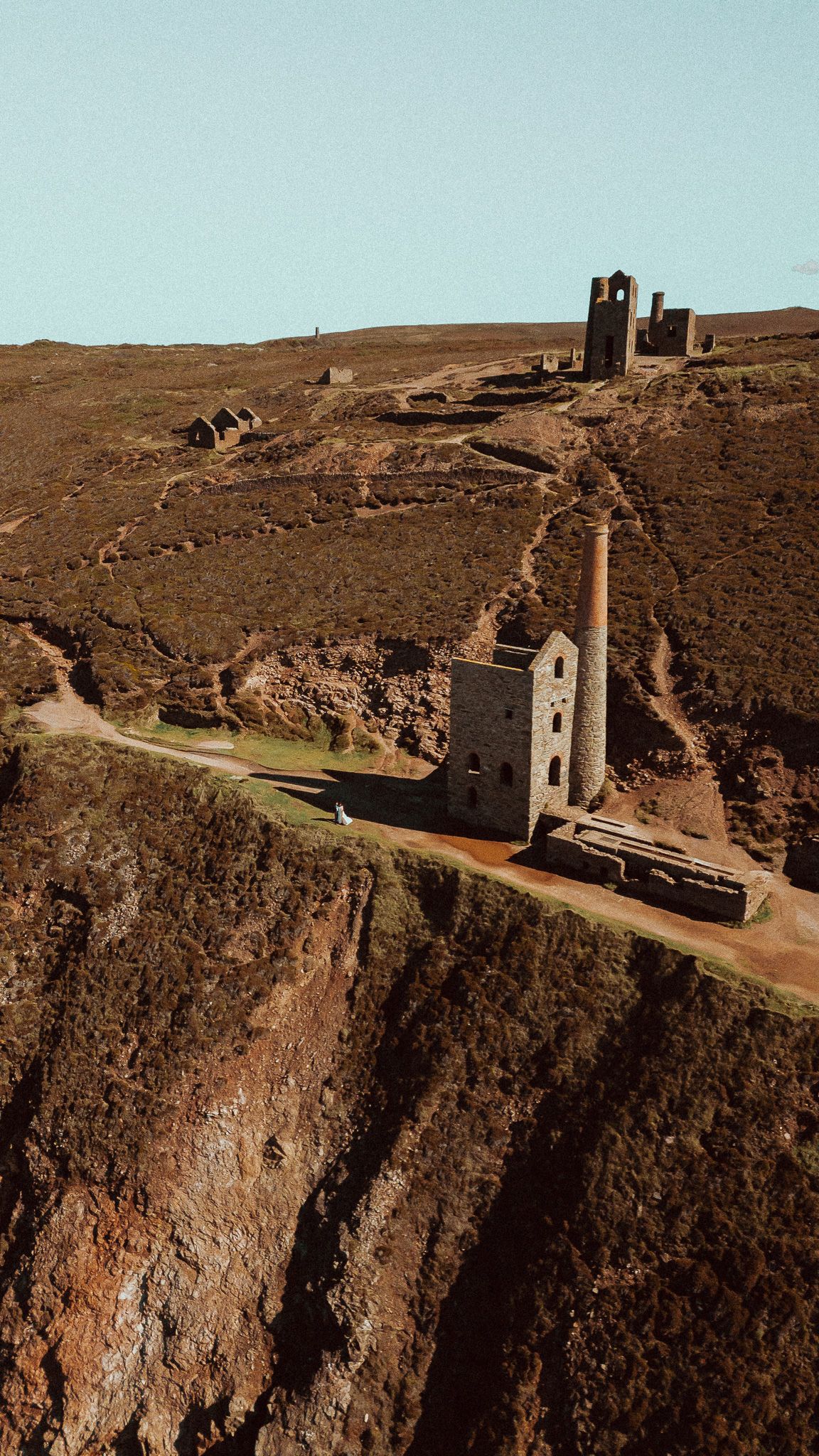 Aerial view of ruined Cornish tin mine engine houses and a tall chimney on rugged heather-covered coastal cliffs under a clear sky.