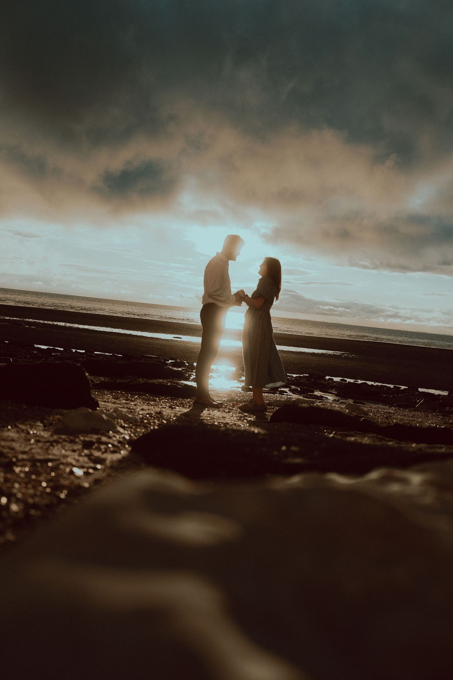 Silhouetted couple holding hands on a rocky beach at sunset, with golden light reflecting on the water beneath a dramatic cloudy sky.