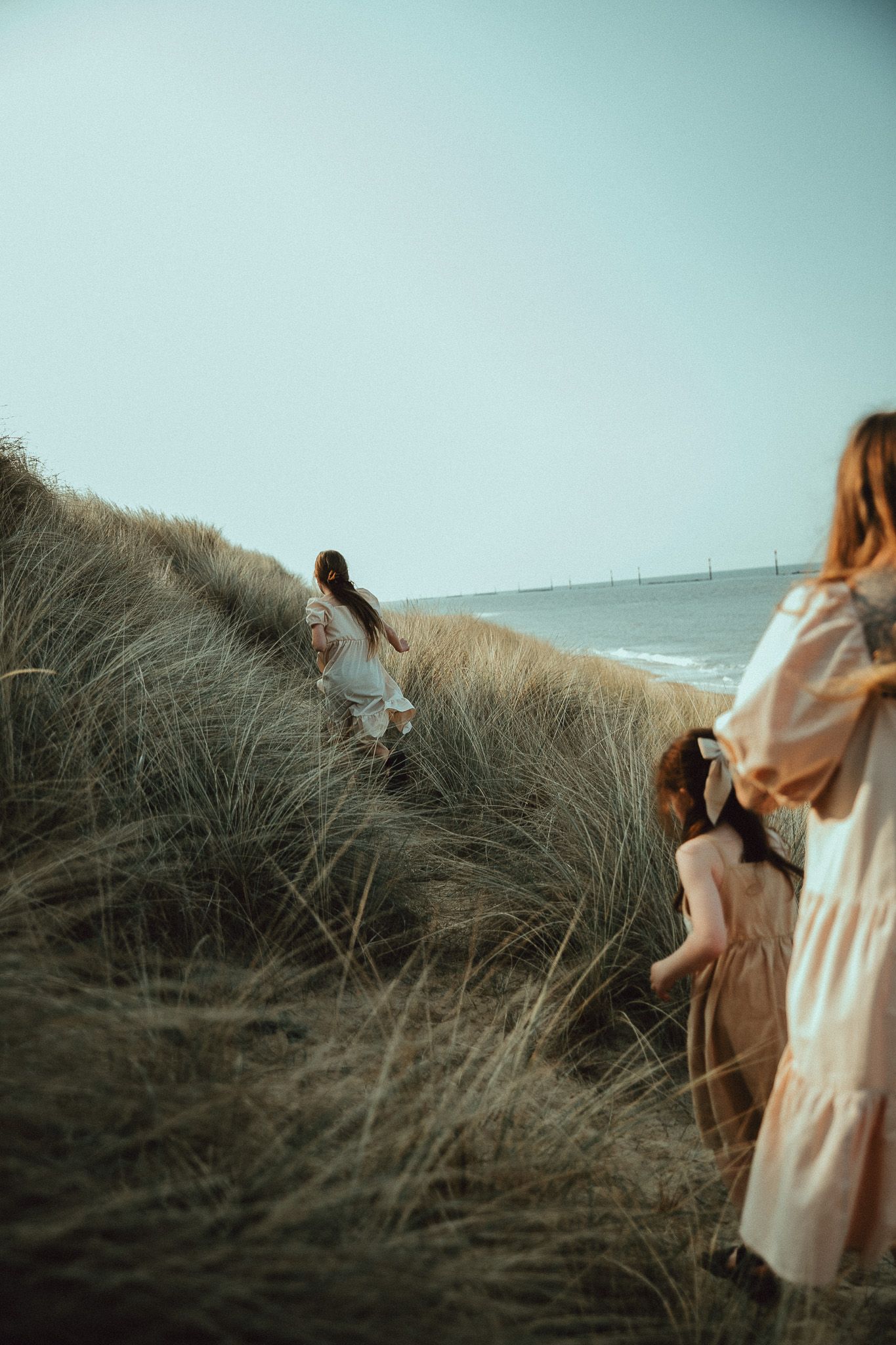 A figure in a flowing dress stands among windswept beach grass on coastal dunes at sunset.