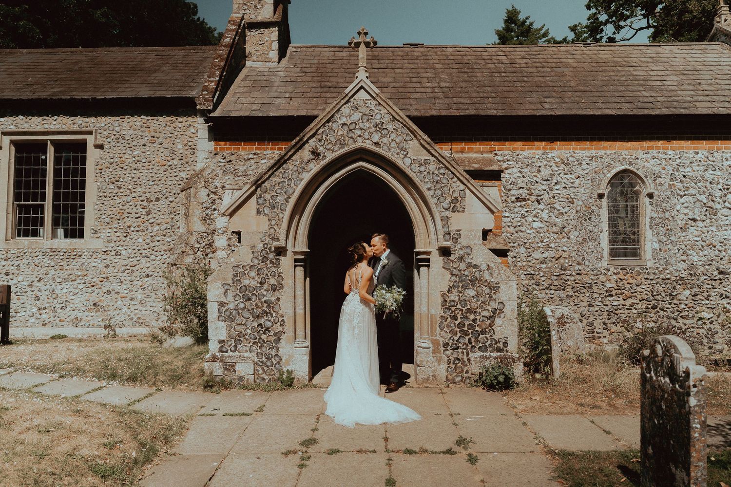 Bride in a white gown and groom in a dark suit embrace in the arched stone doorway of a historic countryside church on their wedding day.