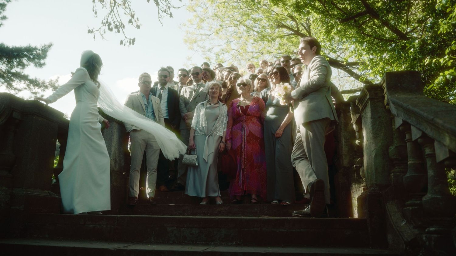 Wedding party poses on stone steps outdoors, bride in white gown with flowing veil among elegantly dressed guests.