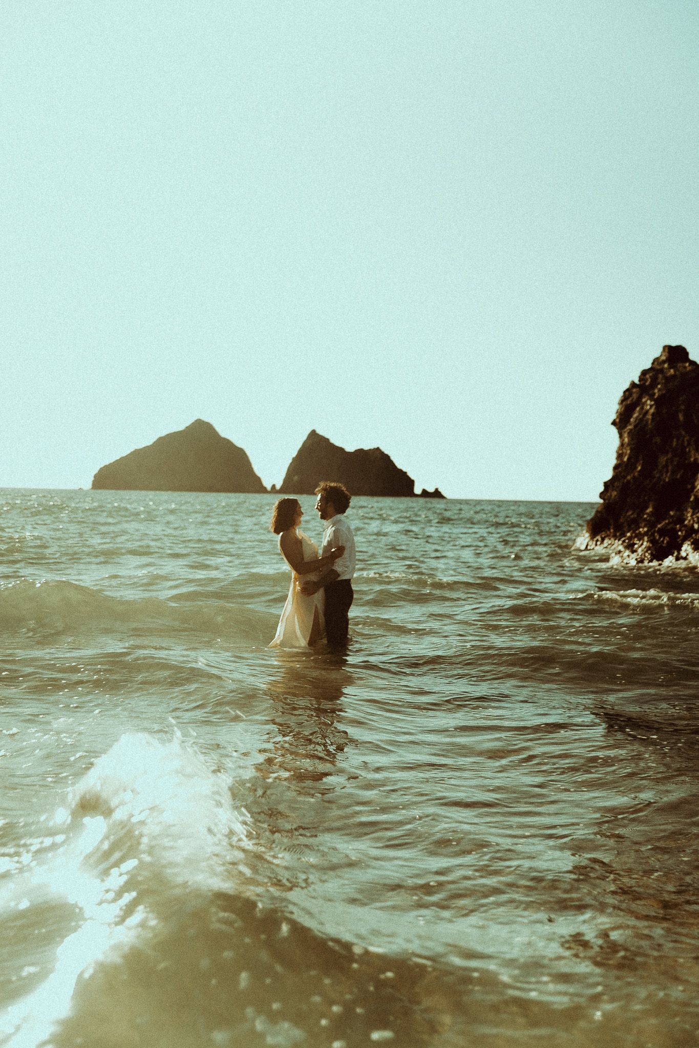 A couple embraces while standing in ocean waves with rocky islands silhouetted against a misty turquoise sky.