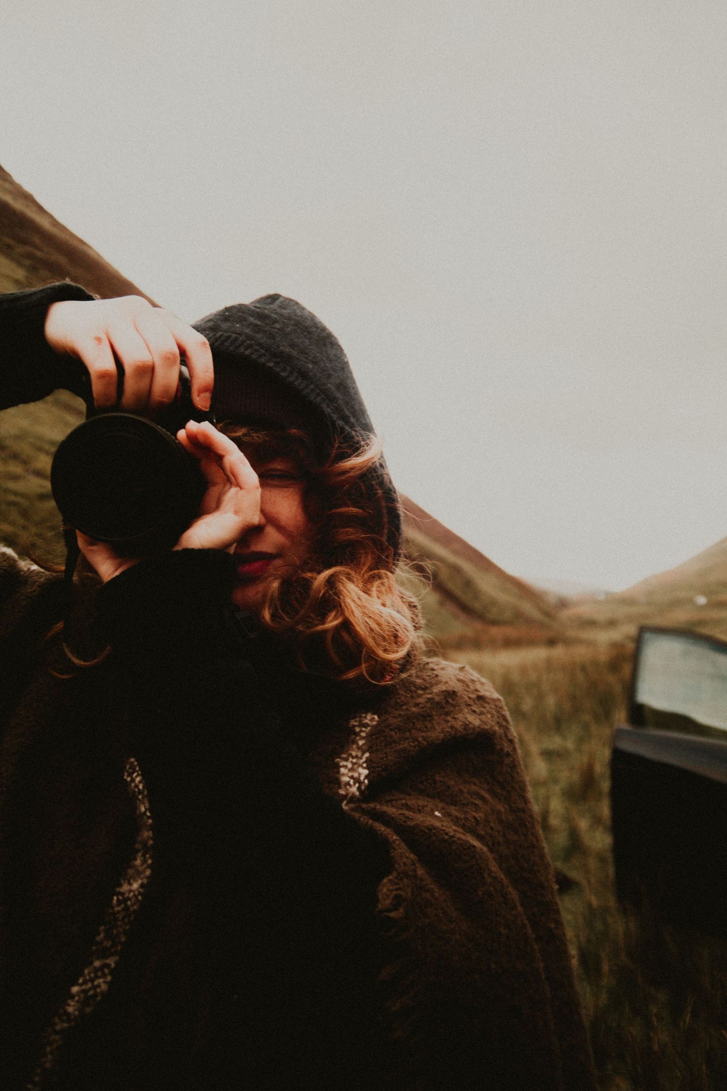 A woman in a dark coat and beanie holds a camera to her face outdoors in a moody, overcast landscape in Scotland.