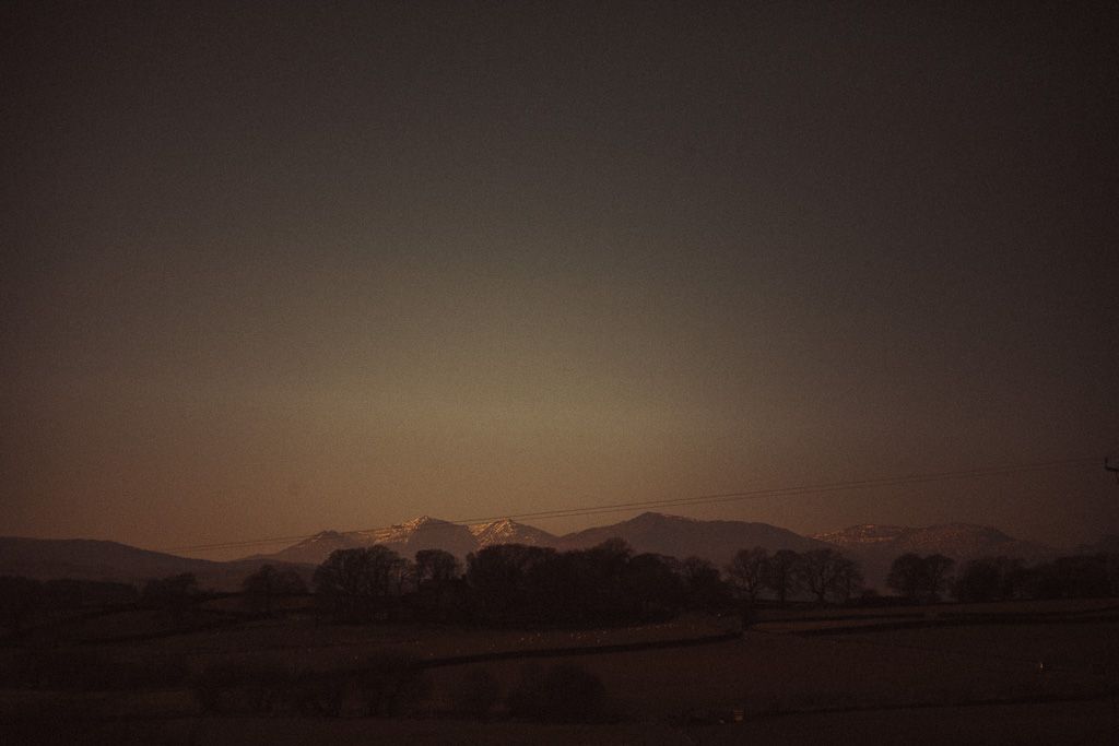 Dark moody landscape at dusk with snow-capped mountains glowing on the horizon under a dim twilight sky in Wales.
