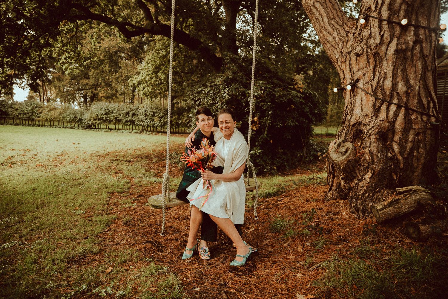 Couple sitting together on a rope swing under a large tree in a lush green park setting.