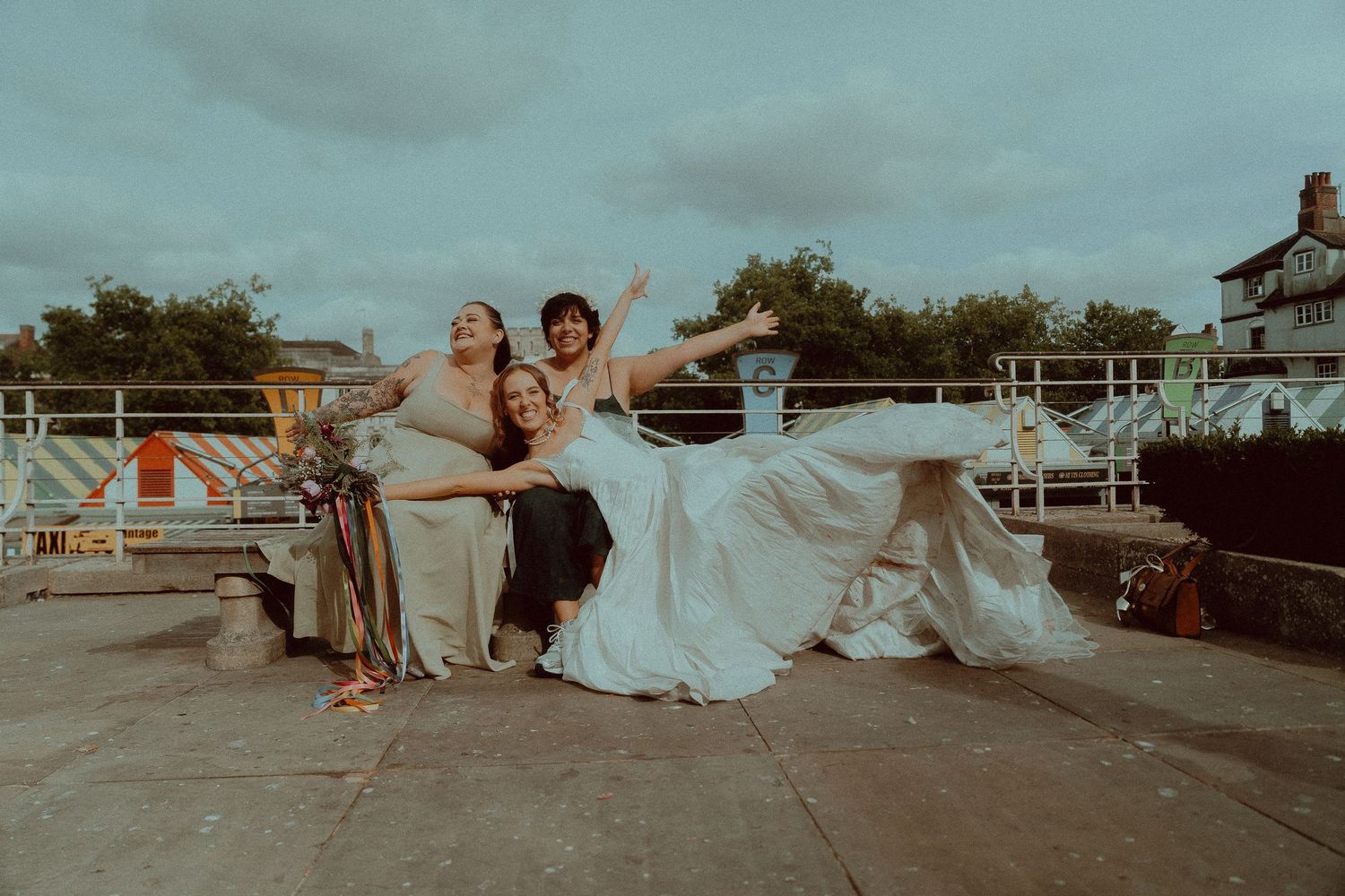 Joyful bride in white gown celebrating with friends outdoors near waterfront on cloudy day.