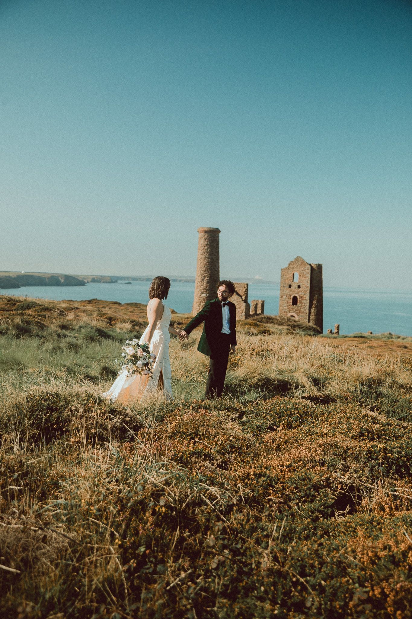A couple walks through a coastal landscape with ancient ruins and wildflowers against a blue ocean backdrop.