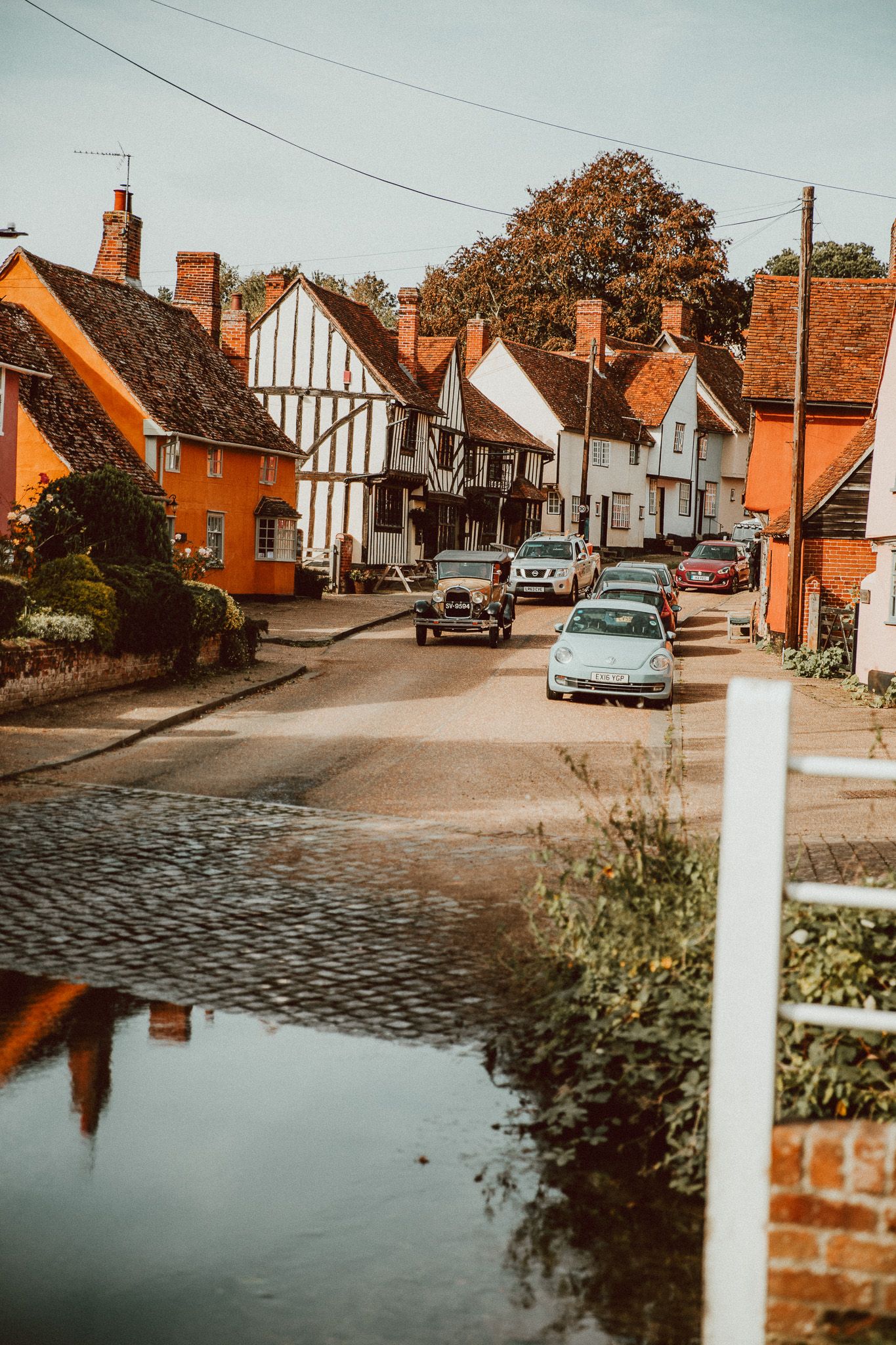 Charming English village street with colorful historic cottages, parked cars, and a reflective puddle in the foreground.