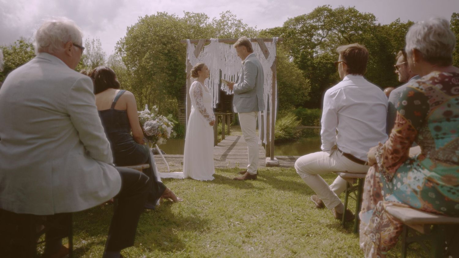 Outdoor wedding ceremony with couple at floral arch, guests seated on chairs in sunny garden setting.
