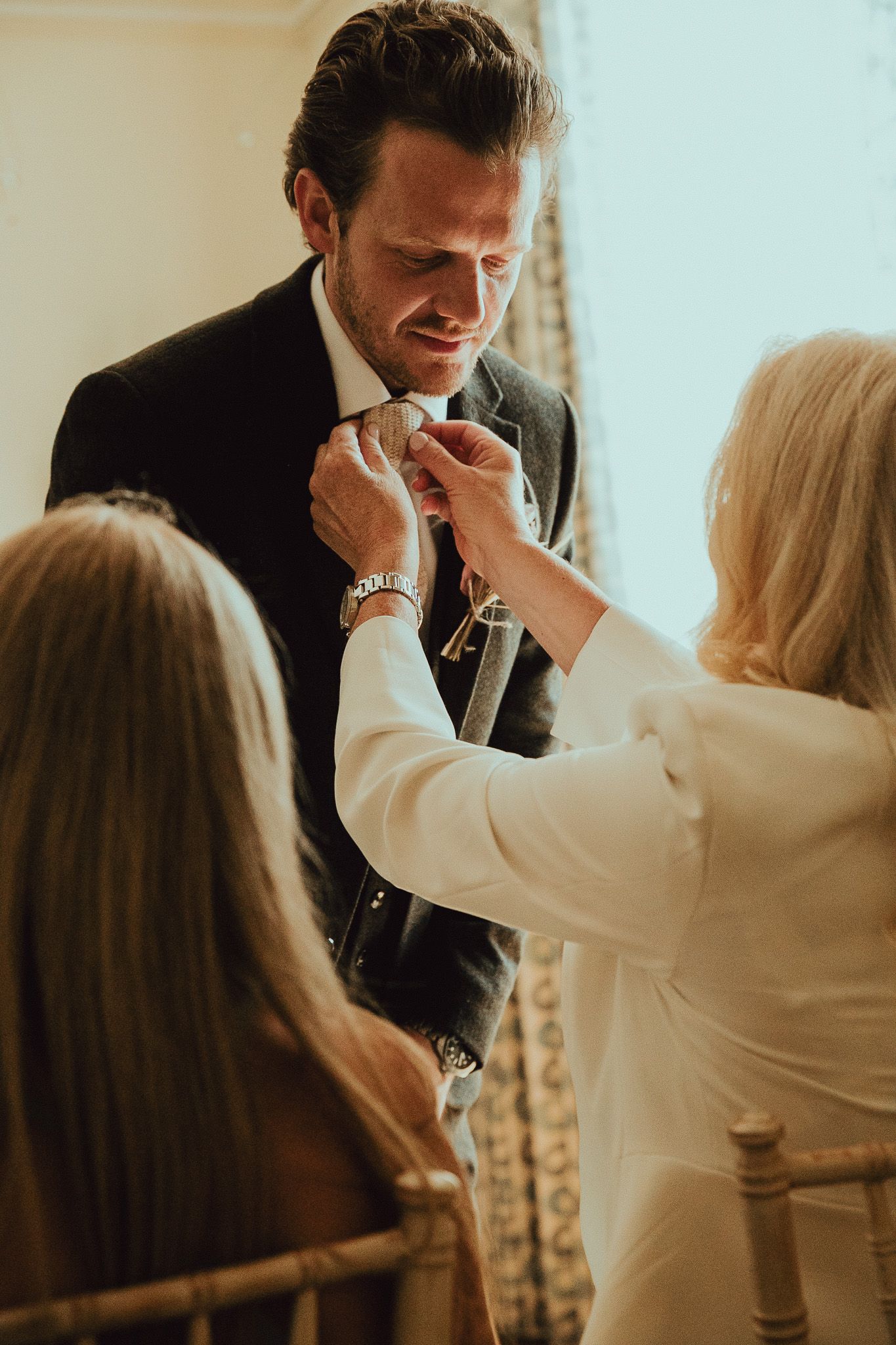 A woman adjusts a groom's tie before his wedding ceremony, assisted by another guest.