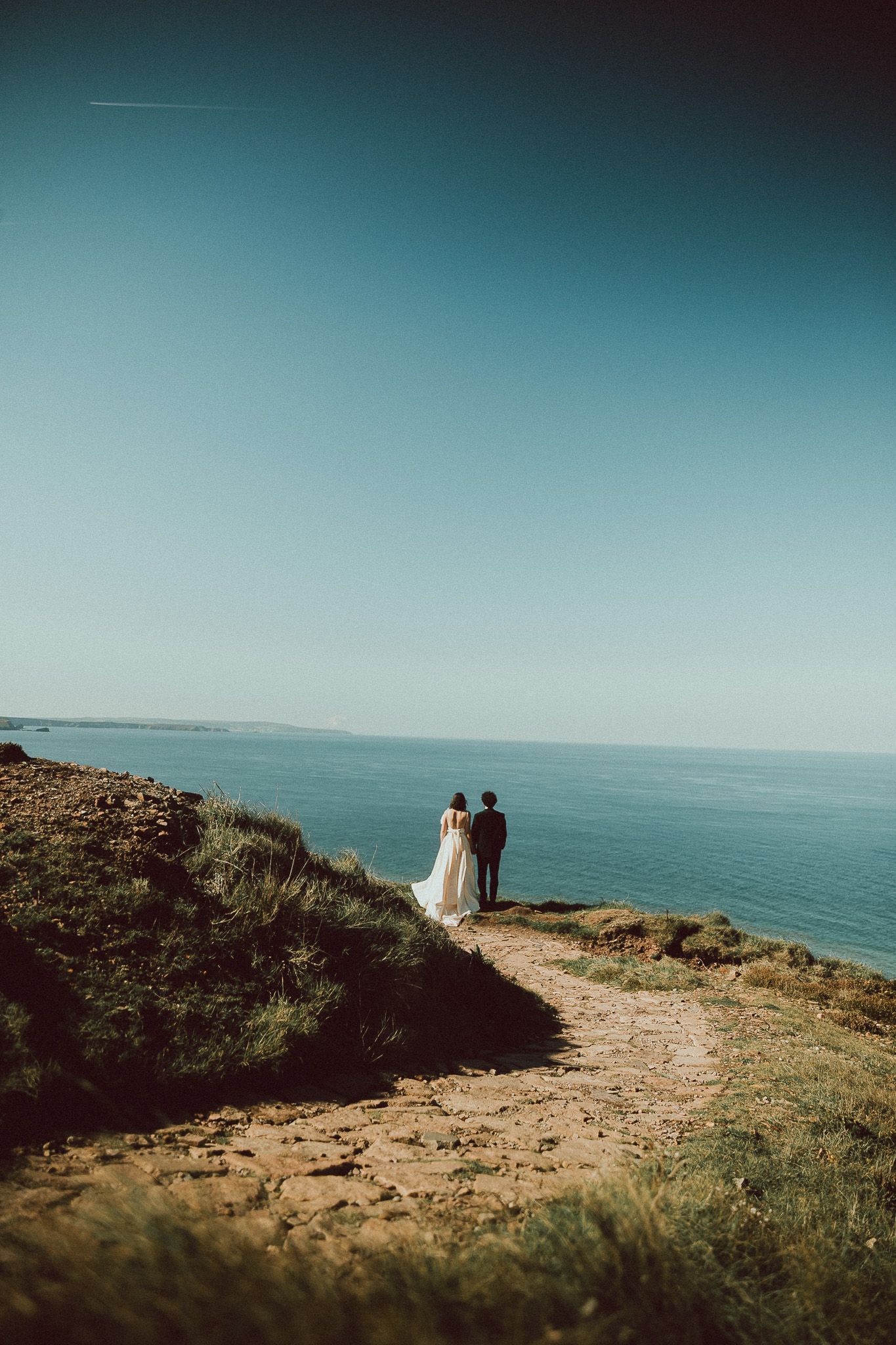 A bride in a white gown and a groom stand together on a grassy coastal cliff path, gazing out at the calm blue sea under a clear sky.