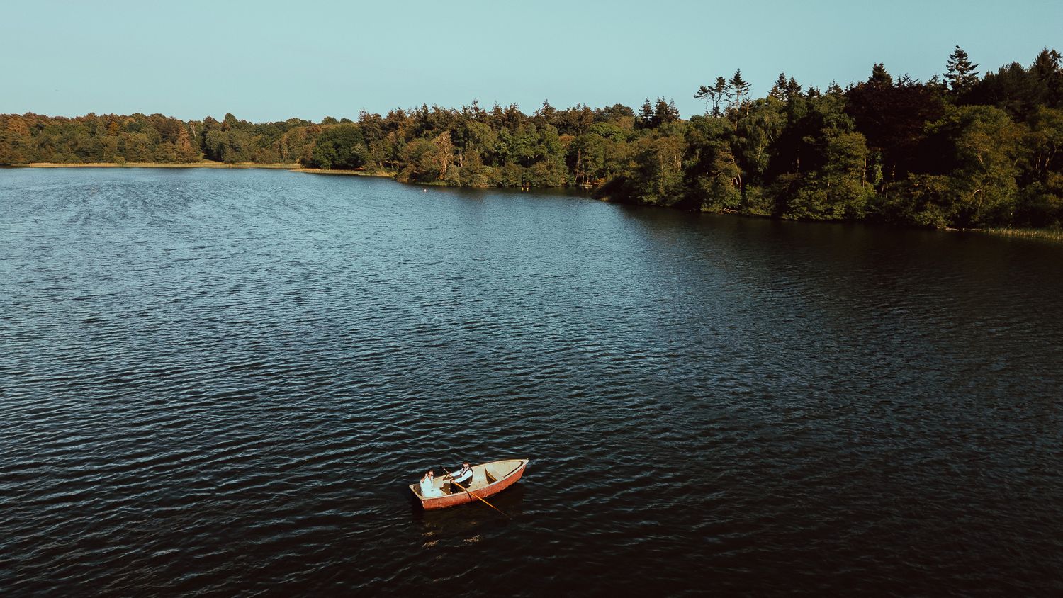 Aerial view of a small boat floating on calm dark lake water, with a forested shoreline and clear blue sky in the background.