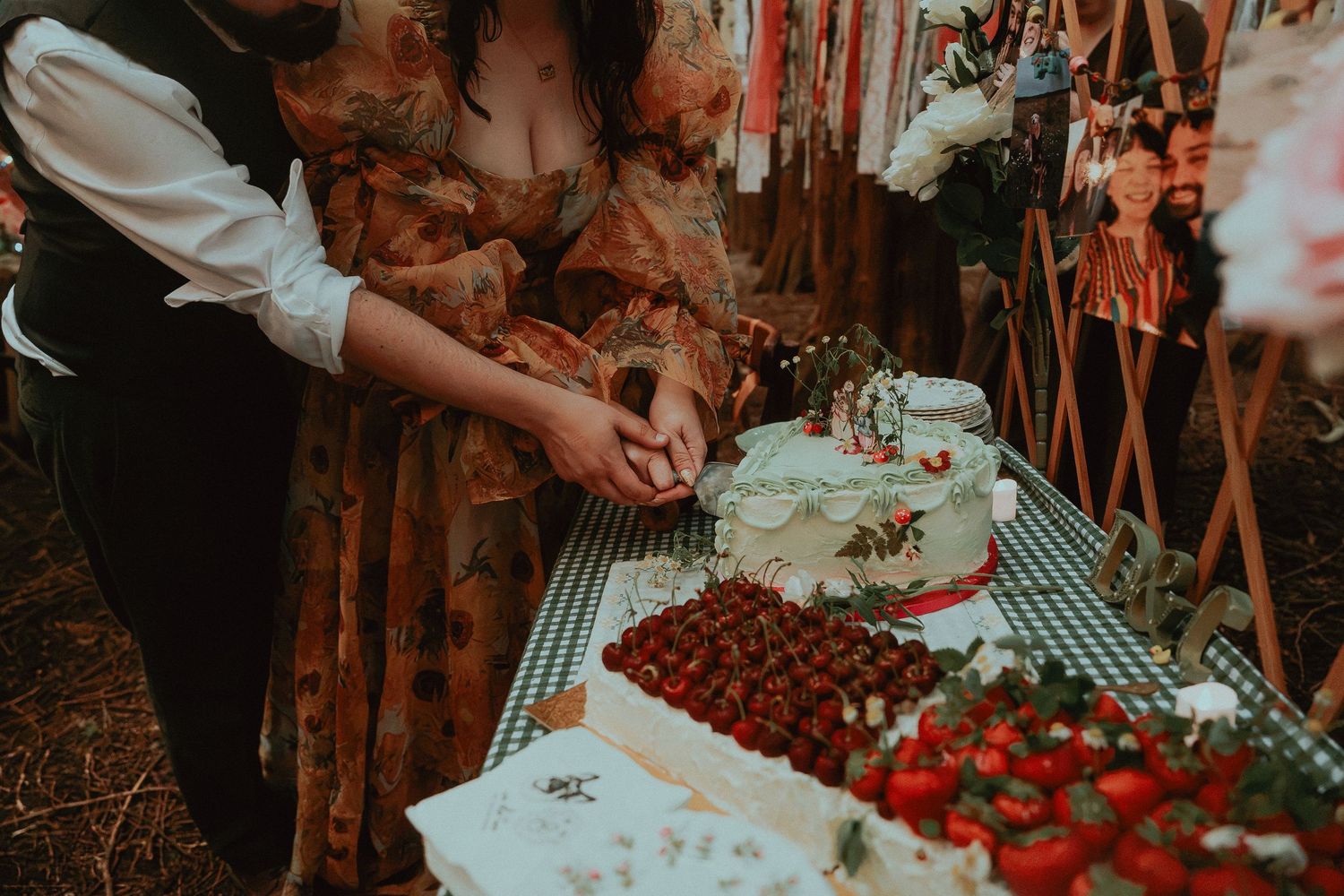 Couple cutting wedding cake at outdoor reception table with fresh strawberries and floral decorations.