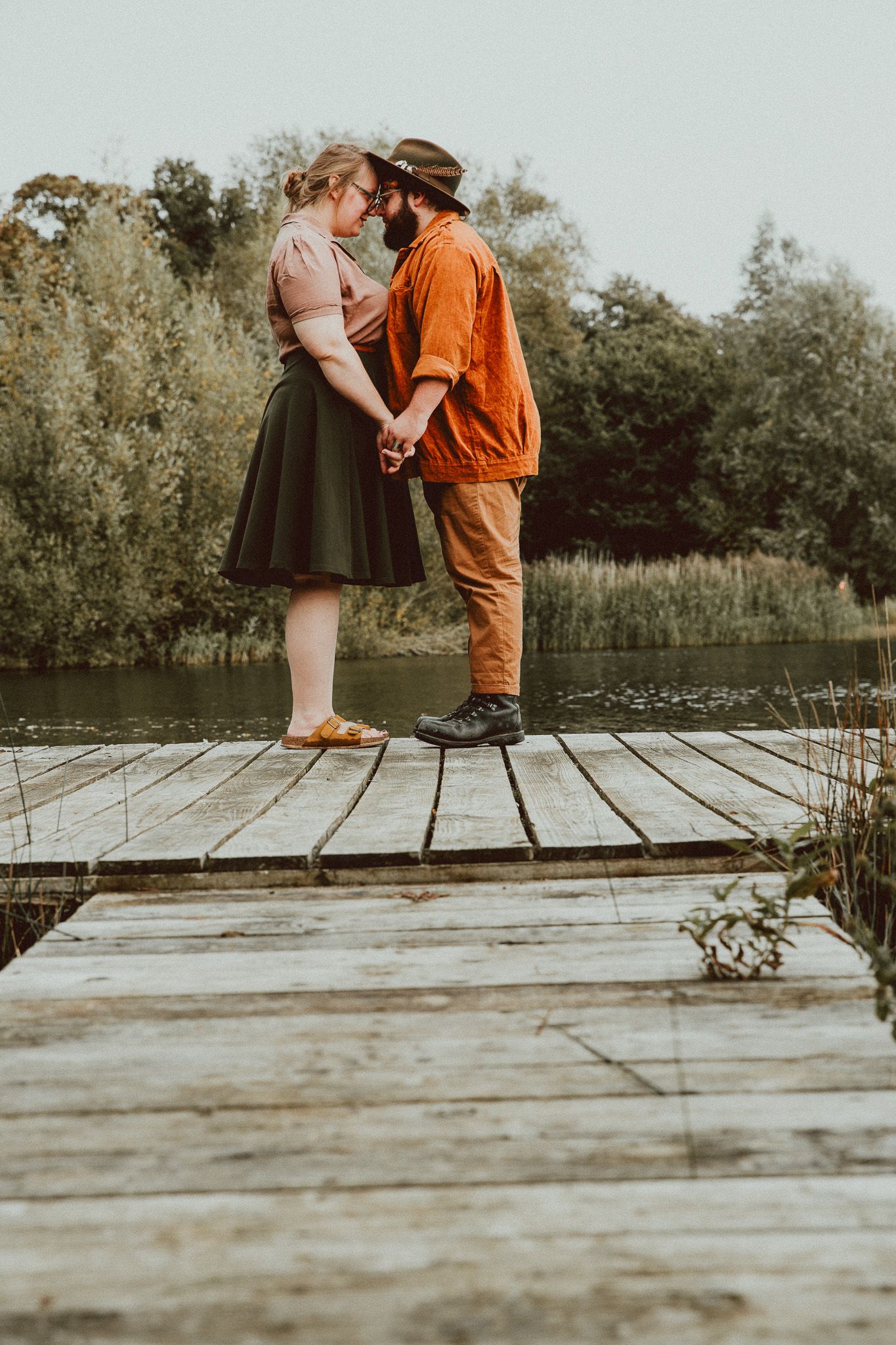 Couple holding hands and kissing on a wooden dock by a serene lake surrounded by lush green trees.