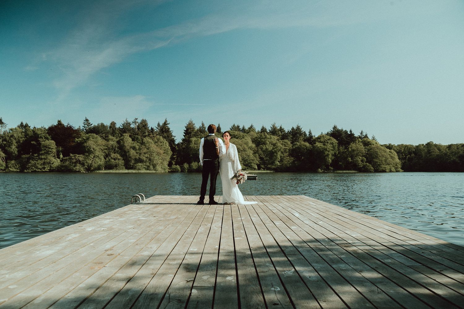 Bride in a white gown and groom in a dark suit embrace on a wooden lake dock, with a calm lake and forested shoreline behind them.