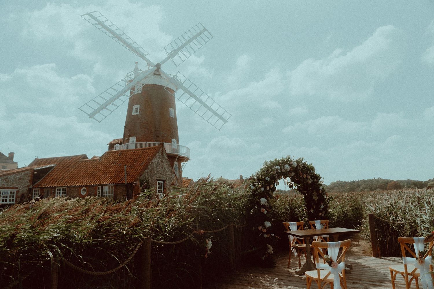 A rustic windmill wedding venue with a floral arch and outdoor seating area under a cloudy sky.