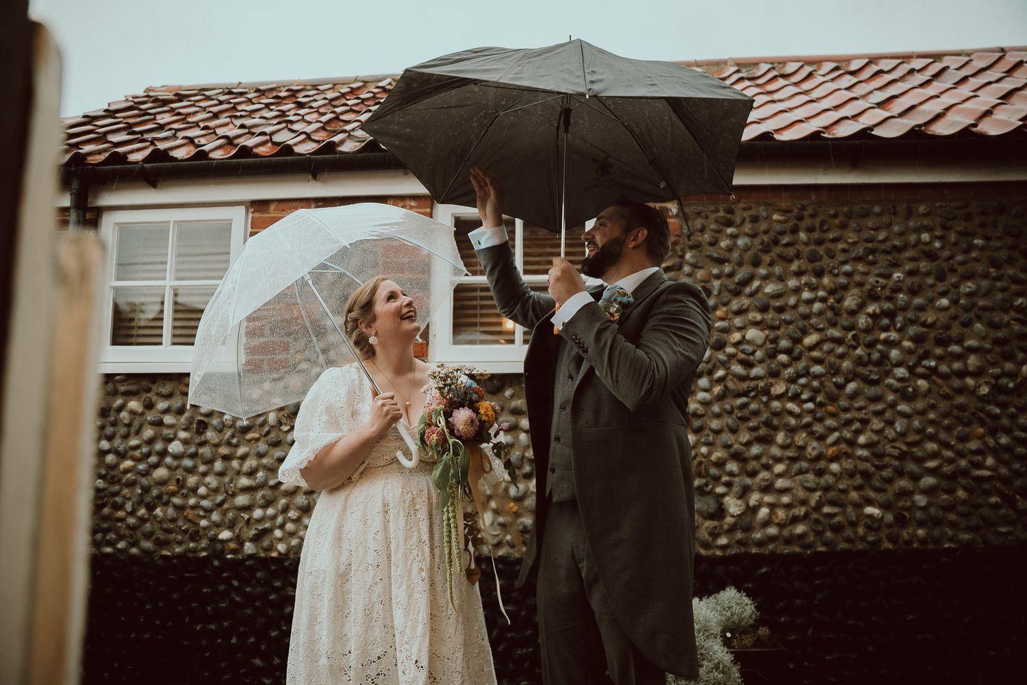 Bride and groom laughing under umbrellas in the rain on their wedding day outside a rustic stone building.