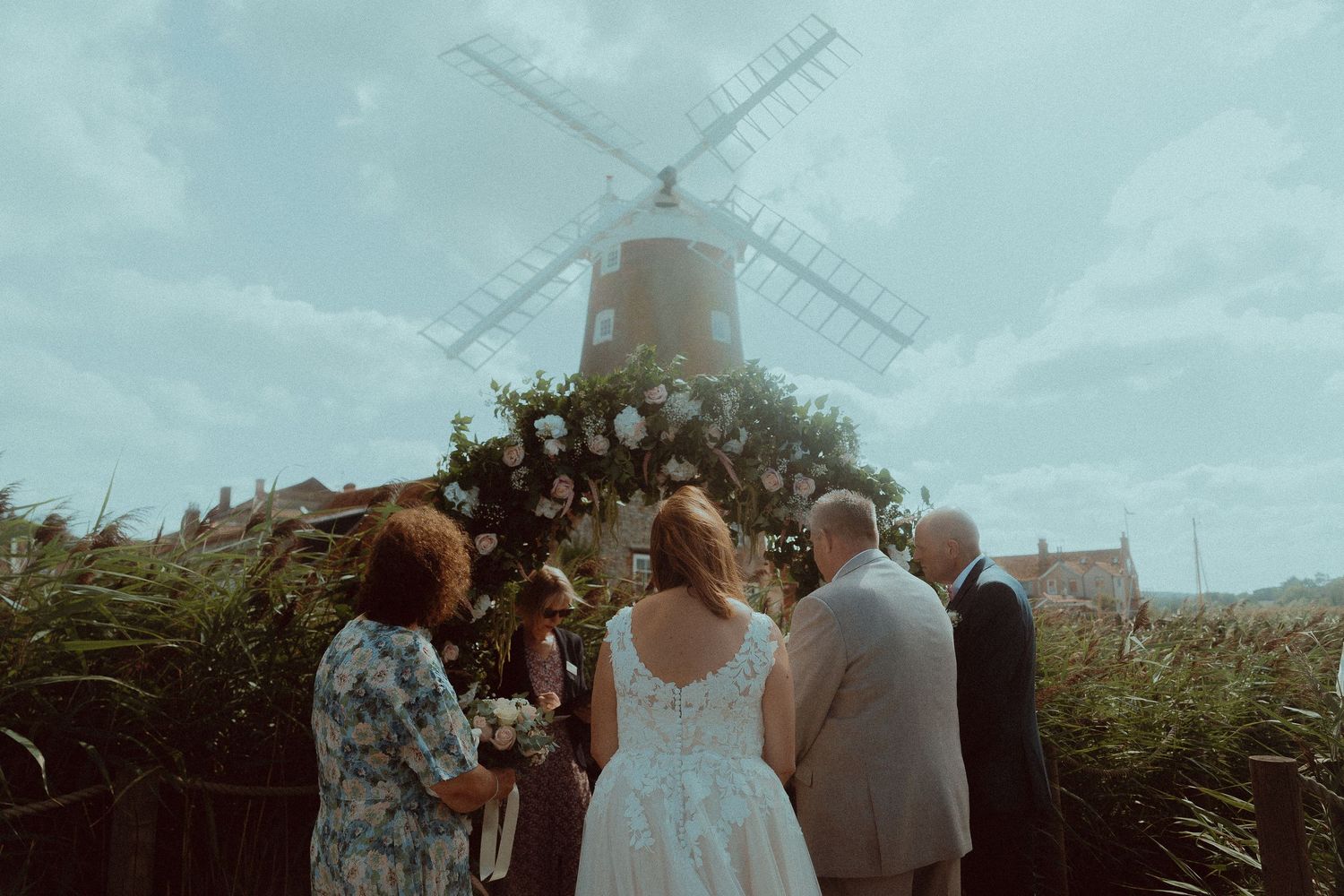 Outdoor wedding ceremony beside a windmill with floral arch, bride and guests facing officiant amid tall grass.