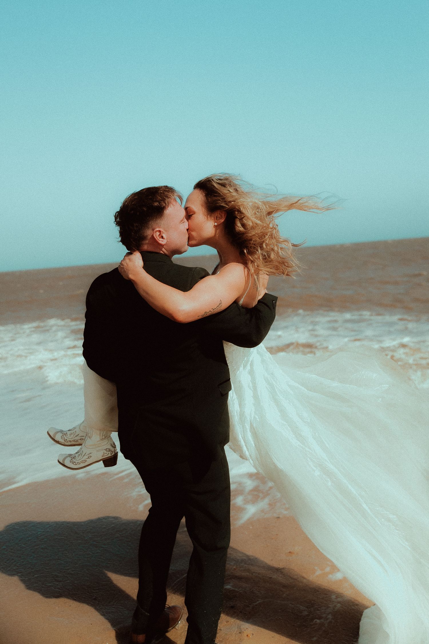 Groom carries bride on beach, sharing a kiss with waves and blue sky behind them, her white dress flowing in the wind.