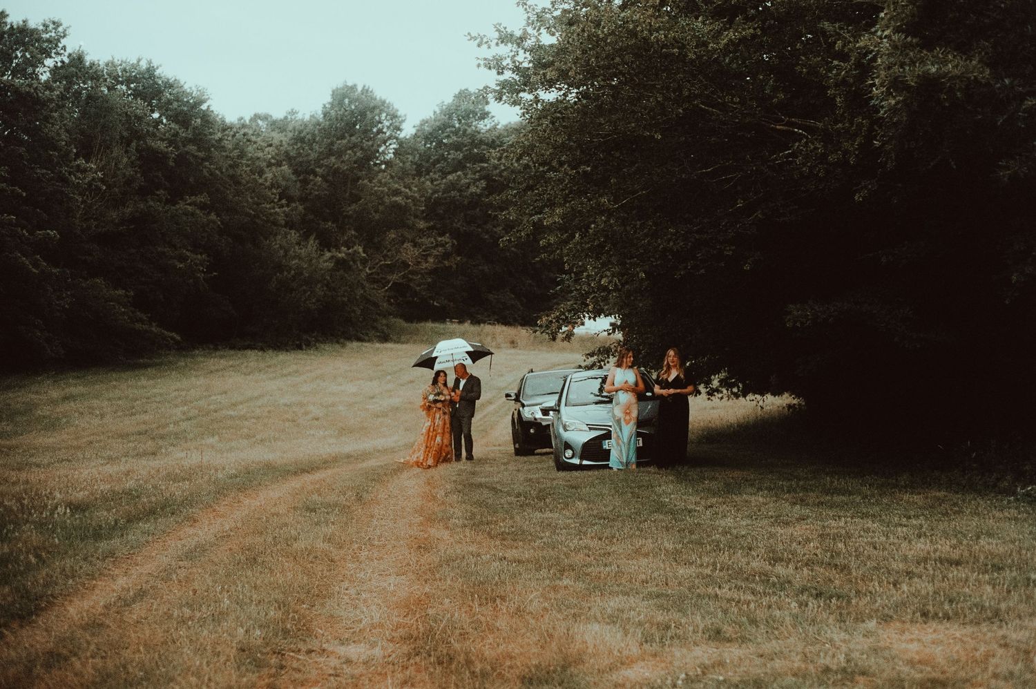 People gathered near a car on a grassy field path surrounded by lush green trees on a sunny day.
