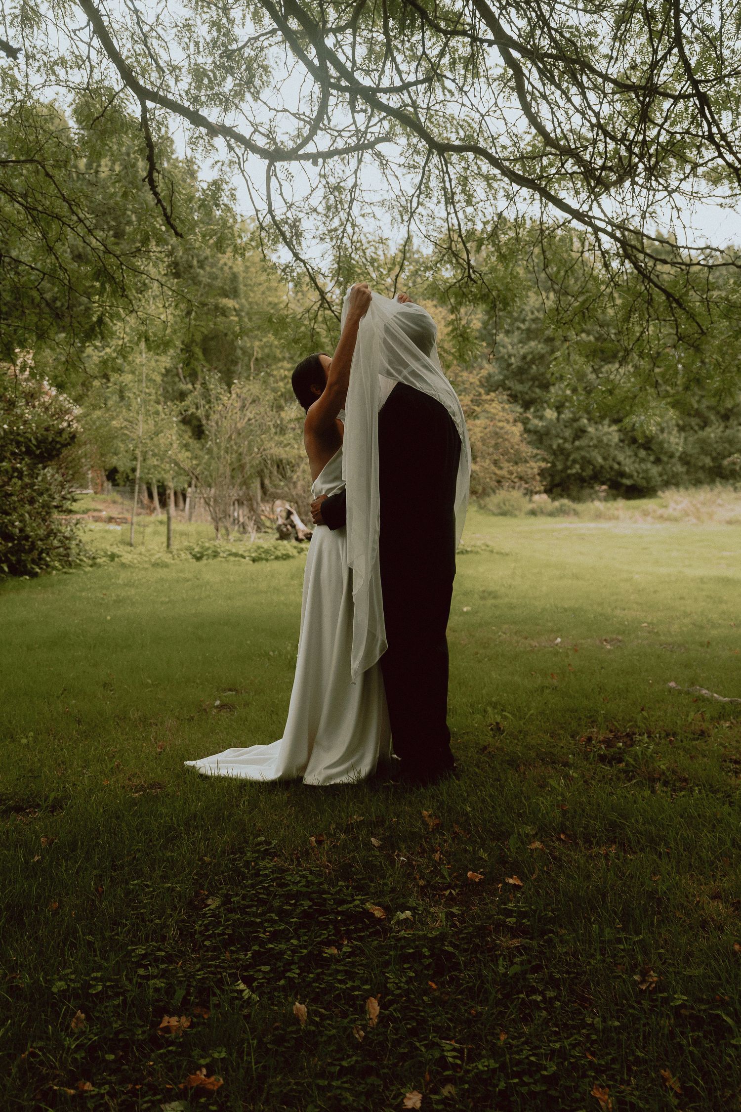 A bride in a white gown and veil embraces her partner in dark attire under a tree in a lush green garden.