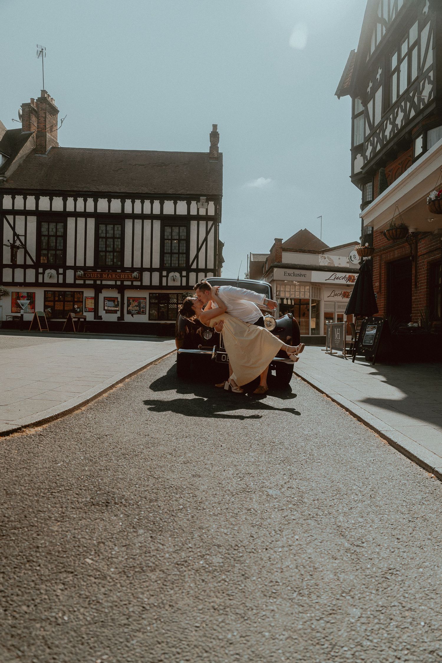 Bride and groom kissing on vintage car in sunny English town square with Tudor-style buildings.